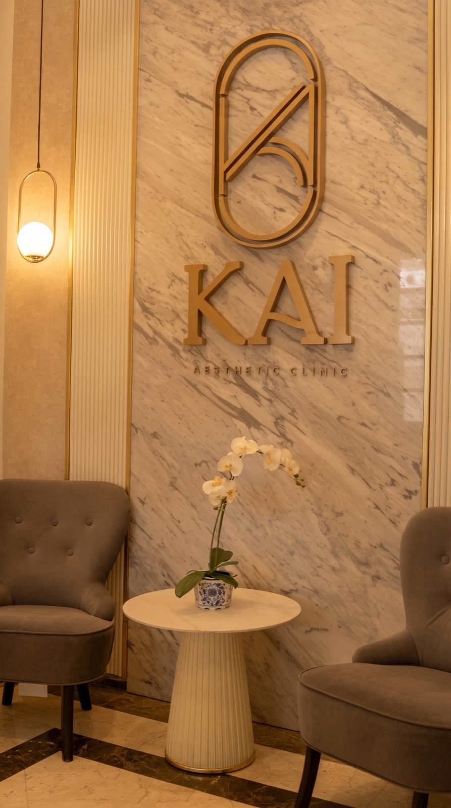 Dinding marmer dengan logo emas KAI Aesthetic Clinic, dua kursi beludru, dan anggrek putih